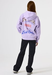 Mujer con cabello largo vistiendo una sudadera lila con el texto "New York Brooklyn East Side", pantalones negros y zapatillas blancas, de espaldas a la cámara.
