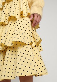 Gonna gialla a balze con pois neri, abbinata a un maglione giallo a maniche lunghe e peloso, mano appoggiata delicatamente sulla gonna.