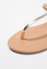 Tan flip-flop sandal med en metallisk guldrem, platt sula och stickad detalj längs kanten. Slät yta på innersulan.
