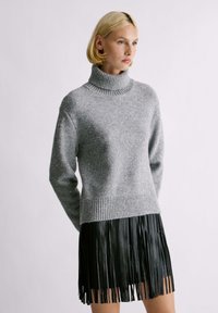 Maglione grigio a collo alto con orlo a costine e accenti glitter, abbinato a una gonna nera plissettata con frange, mostrato su uno sfondo neutro.