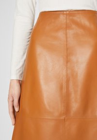 Femme portant une jupe en cuir marron taille haute avec des détails de coutures verticales, associée à un haut blanc côtelé à manches longues.
