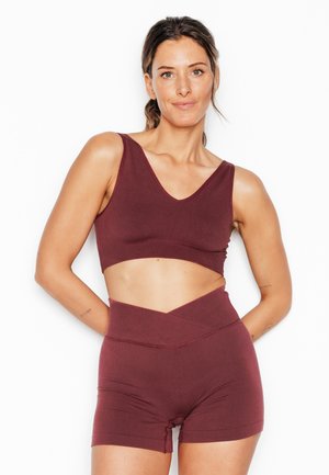 Femme debout, les mains derrière le dos, portant un brassard de sport bordeaux et un short taille haute sur fond blanc.