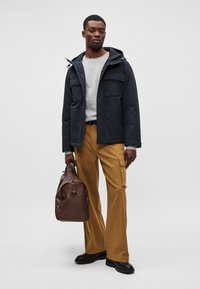 Casaco de padronagem da marinha com capuz e múltiplos bolsos, sweatshirt cinza por baixo, calças cargo bege, bolsa de couro castanho, botas pretas.