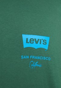 Zielona bawełniana koszulka z niebieskim logo Levi's, napisem "SAN FRANCISCO" oraz "California" w stylu kursywy, na tle gładkiej faktury tkaniny.