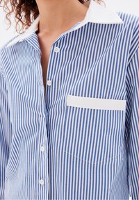 Camicia a righe con bottoni, caratterizzata da righe verticali blu e bianche, colletto bianco e taschino sul petto con finiture curate. Tessuto morbido.