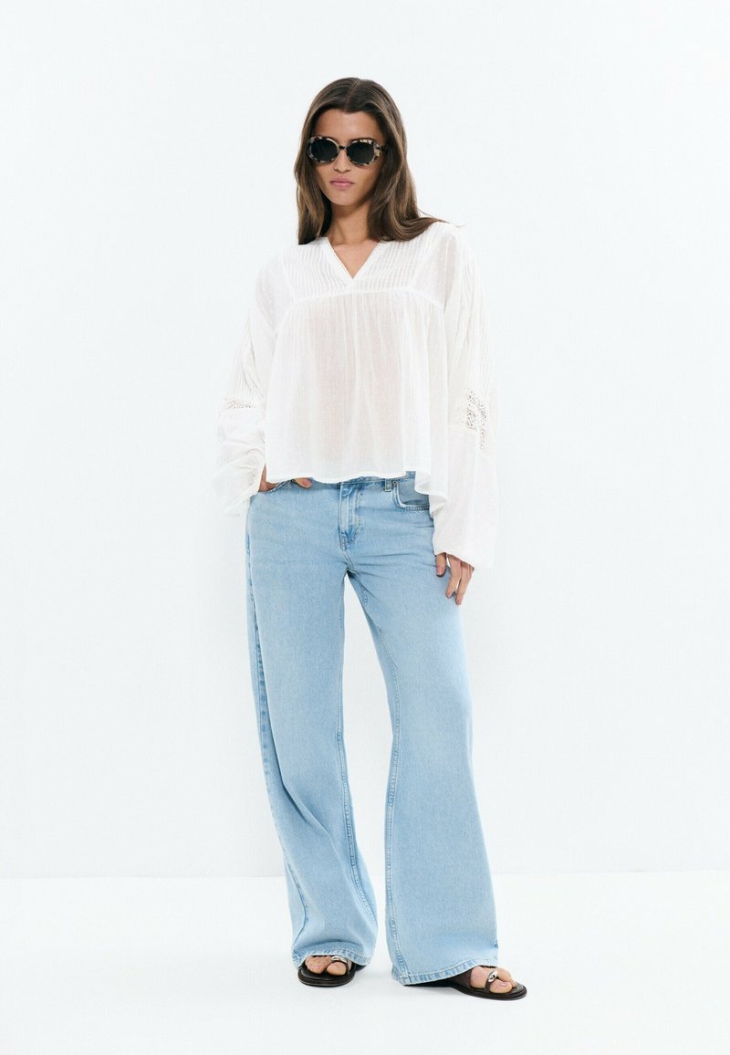 Mujer con blusa blanca de gran tamaño, vaqueros de pierna ancha azul claro, sandalias negras y gafas de sol oscuras, de pie contra un fondo blanco liso.