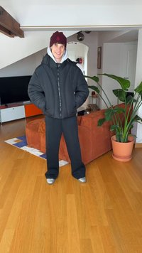 Melna pūkaina jaka ar rāvējslēdzēja aizdari, zem tās pelēks kapucis, melnas bikses un sudraba krāsas apavi. Aksesuāri: sarkanbrūna cepure.