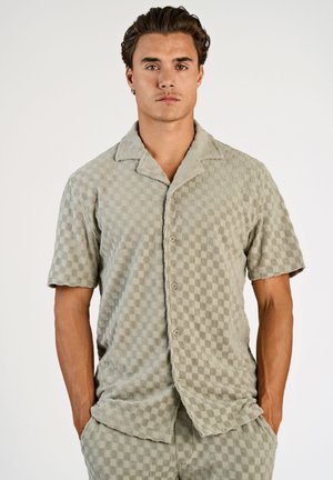 Uomo che indossa una camicia a maniche corte color grigio chiaro con bottoni e pantaloncini abbinati con un delicato motivo a quadretti, con le mani nelle tasche.