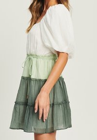 Vestido ligero en capas con mangas abullonadas. El corpiño blanco se transforma en degradados de verde. Presenta una cintura con cordón y una tela texturizada.