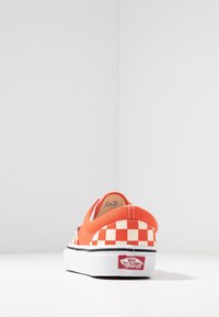 Rød lærredssneaker med hvid ternet mønster på hælen, hvid gummisål og sort stribe; slip-on design med logo bagpå.