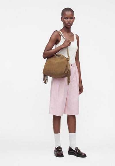 Femme en débardeur blanc, short rose clair longueur genou, chaussettes blanches, mocassins foncés, tenant un sac à main en daim marron avec détail à franges sur l’épaule.