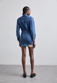 Denim mini haljina s dugim rukavima, strukturiranim krojem, svijetloplave boje i kontrastnim detaljem oznake na leđima. Gumbi na manžetama rukava.