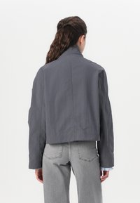 Kuttet grå jakke med en glatt tekstur og høy krage, med et enkelt design og litt oversized ermer. Kombinert med lysegrå jeans.