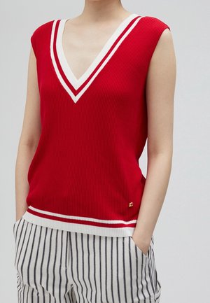 Haut en maille rouge sans manches avec des rayures blanches au niveau du col en V et de l'ourlet, porté avec un pantalon blanc à fines rayures noires verticales.