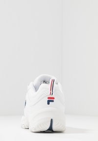 Baskets de sport blanches à semelle épaisse vues de dos, avec des détails rouges et bleu marine et une languette à rayures rouges et bleu marine.