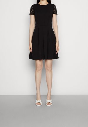 Vrouw die een zwarte jurk met korte kanten mouwen en witte sandalen met open tenen draagt, staand tegen een effen lichte achtergrond.