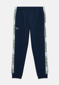 Pantaloni de trening bleumarin cu talie elastică, având dungi laterale albe și verzi cu branding "LACOSTE" și un logo cu crocodil.