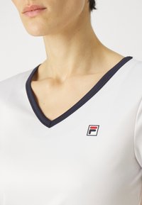 Bílý sportovní top s námořnickým V-výstriem a malým červeným a černým logem na levé části hrudi. Hladká, lehká tkanina.