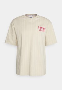 Béžové tričko s krátkým rukávem, bílé vertikální pruhy a červené logo "TOMMY JEANS" na levé hrudi. Bavlněný materiál, volný střih.
