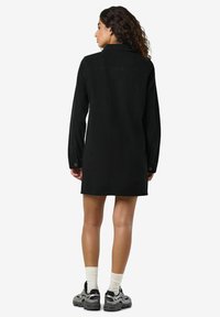 Žena s kudrnatými vlasy nosí černé krátké šaty s dlouhým rukávem, bílé ponožky do půli lýtek a šedé černé tenisky, stojící na bílém pozadí.