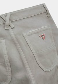 Pantaloni corti in denim beige con una tasca posteriore scucita. Presentano un'etichetta triangolare rossa e nera con il logo "Guess" e cuciture piatte.