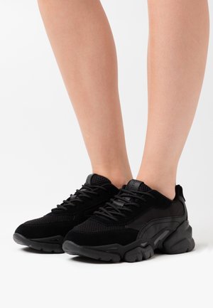 Person wearing black chunky sneakers with mesh and suede details, standing on a white surface.