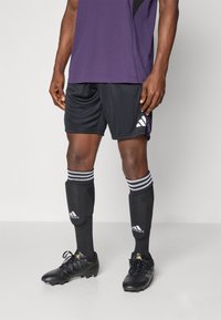 Sorte fodboldshorts, lilla trøje, sorte knæstrømper med hvide striber og sorte fodboldstøvler med guld accenter. Adidas branding synlig.