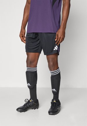 Zwarte voetbalkorte broek, paarse jersey, zwarte kniebeschermers met witte strepen, en zwarte voetbalschoenen met gouden accenten. Adidas-logo zichtbaar.