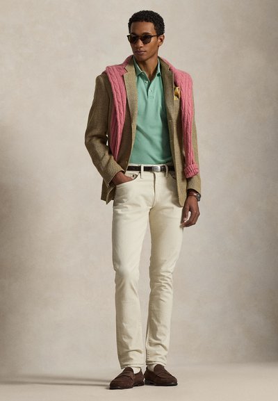 Moški z sončnimi očali, bež suknjičem, mint polo majico, skoraj belimi hlačami, rjavimi mokasini in roza puloverjem, ki je vržen čez ramena, stoji pred enobarvnim ozadjem.