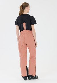Roze skibroek met verstelbare zwarte banden, elastische tailleband en gecompartimenteerde onderbenen. Draagt over een zwart T-shirt en zwarte skir boots.