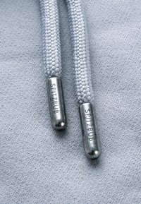 Gros plan sur les cordons de capuche gris clair terminés par des embouts en argent avec "SPITZBUB" gravé sur les embouts métalliques, sur un fond de tissu gris doux.