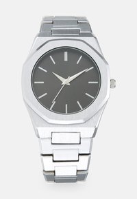Sølv analog ur med et oktagonalt hus, grå urskive, sølvfarvede timeindikatorer og et armbånd af rustfrit stål. Minimalistisk design med en glat tekstur.