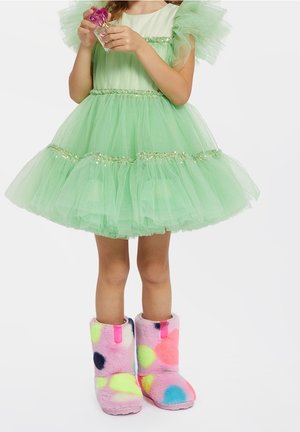 Enfant portant une robe en tulle verte avec des bordures dorées et des bottes colorées et duveteuses, tenant un petit flacon de parfum à capuchon rose.