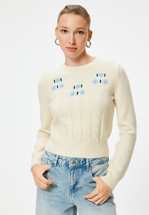 Cremfarbener Kabelstrickpullover mit blauer Blumenstickerei und grünen Akzenten, mit Rundhalsausschnitt und gerippten Bündchen, kombiniert mit blauen Jeans.