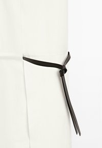 White garment with a tailored fit featuring a black leather tie detail at the side, adding contrast and texture to the design.