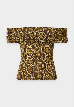 Kvinde iført en gul og brun ormeprints off-shoulder jumpsuit med sorte sko, stående mod en ensfarvet hvid baggrund.