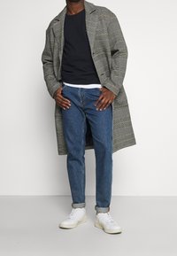 Langer Karomantel in Grau und Schwarz über einem navyblauen Sweater, kombiniert mit aufgerollten Denim-Jeans und weißen Sneakers. Details umfassen ein karierter Muster.