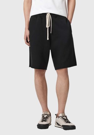 Uomo che indossa pantaloncini neri larghi fino al ginocchio con coulisse beige, maglietta bianca e sneaker grigio chiaro con suole e lacci neri.