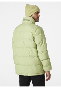 Zelená puffová bunda s prošívaným designem, s vysokým límcem a nápisem "Helly Hansen" na zadní straně. Spárovaná se světle žlutou čepicí.
