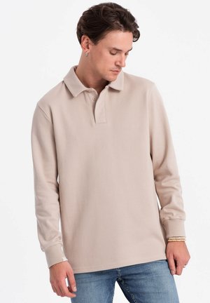 Beige Langarm-Polohemd mit klassischem Kragen, strukturiertem Stoff und geradem Saum, kombiniert mit blauen Jeans. Minimale Details.