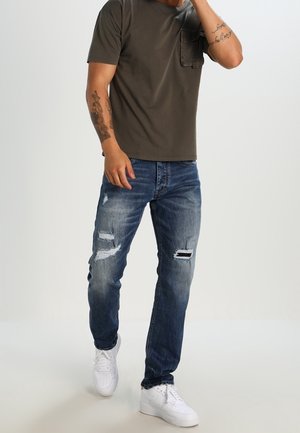 Olijfgroene t-shirt met korte mouwen en een zak, gecombineerd met versleten blauwe jeans met scheuren, en witte sneakers.
