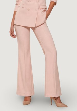 Donna che indossa pantaloni svasati su misura color rosa chiaro, blazer doppiopetto abbinato e scarpe beige con tacco alto, in piedi con una mano in tasca.