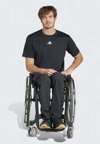 Zwarte sportshirt met wit logo, gecombineerd met donkere broek en zwarte schoenen. Persoon zittend in een rolstoel. Eenvoudig, functioneel ontwerp.