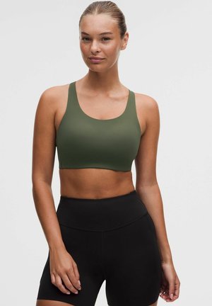 Donna con pelle chiara e capelli tirati indietro indossa un reggiseno sportivo verde oliva e pantaloncini neri a vita alta, in piedi contro uno sfondo chiaro e uniforme.