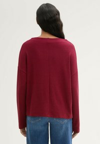 Burgunderfarbener Strickpullover mit weitem Ausschnitt, überschnittenen Schultern und lockerer Passform. Rückennaht-Detail ist sichtbar. Kombiniert mit blauen Jeans.