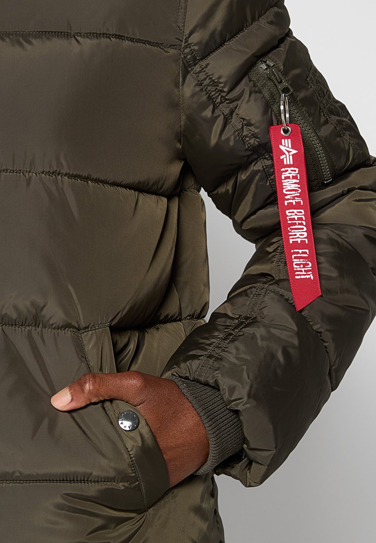 Chaqueta tipo abrigo en verde oliva con textura acolchada, bolsillo con cremallera en la manga y una etiqueta roja que dice "RETIRAR ANTES DEL VUELO."