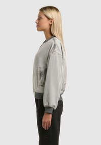 Femme aux cheveux blonds longs portant une veste bomber gris clair et un pantalon noir, debout de profil devant un fond uni.