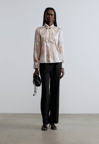 Světle růžová halenka s límečkem na zavázání a květinovým vzorem, doplněná černými kalhotami s širokými nohavicemi. Černá kabelka dodává outfitu kontrast.