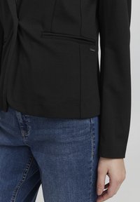 Černé přiléhavé sako se strukturovaným designem, jednou kapsou a hladkým materiálem. V kombinaci s modrými džíny; měkká textura a klasický střih.