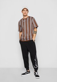 Brunstribet kortærmet skjorte med blå striber, sorte sweatpants med hvidt design og sorte sneakers med hvide accenter.